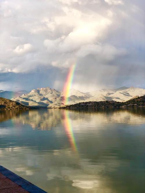 A serene lake reflects mountains and a vibrant rainbow under a cloudy sky.