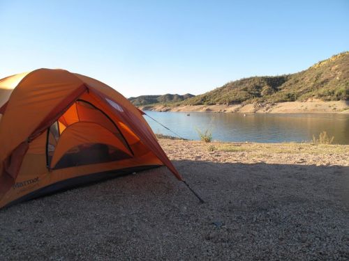 A bright orange tent set up by a calm lake, surrounded by hills and clear blue skies.