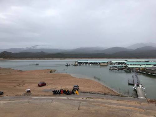 A cloudy view of a lake with boats, a dock, and sandy shores, surrounded by mountains in the background.