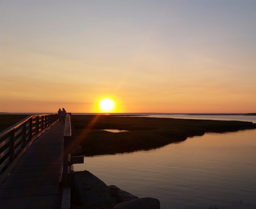 A serene sunset over a calm waterway, with a wooden pier and silhouettes of people enjoying the view.