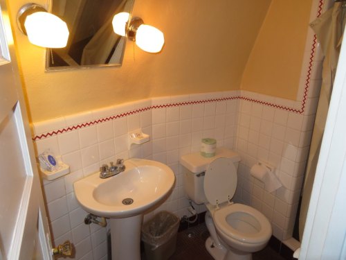 A small bathroom featuring a toilet, sink, mirror, and light fixtures, with tiled walls and a shower curtain.