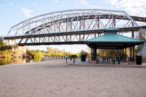 A sandy area with a gazebo and a bridge overhead, surrounded by trees and water.