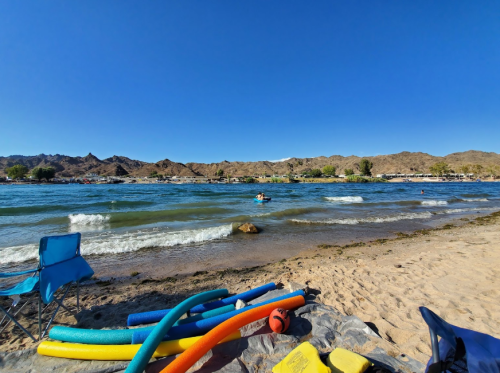 A sunny beach scene with colorful pool noodles, gentle waves, and mountains in the background.