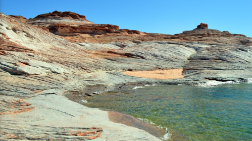 Rocky shoreline with smooth, layered formations and clear blue water under a bright blue sky.