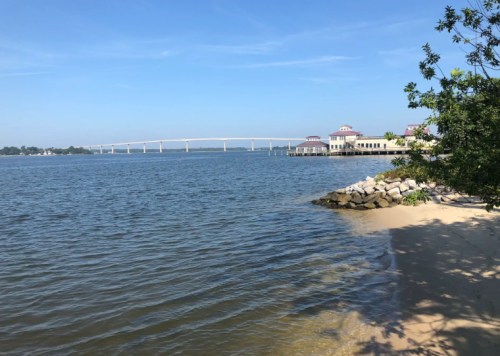 A serene waterfront view featuring a bridge, sandy shore, and buildings under a clear blue sky.