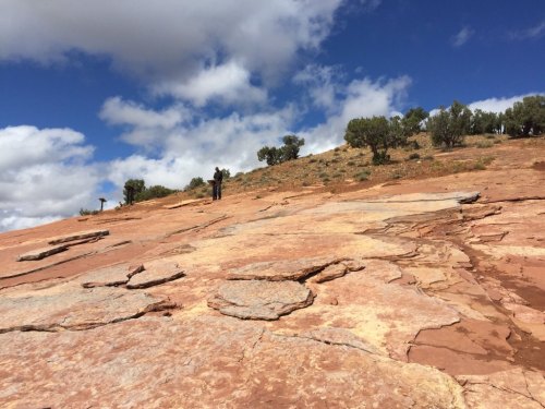 A rocky landscape with two people walking on a sloped surface under a partly cloudy sky.