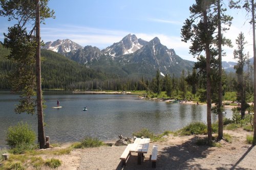 A serene lake surrounded by mountains, with people kayaking and a picnic table in the foreground under a clear blue sky.