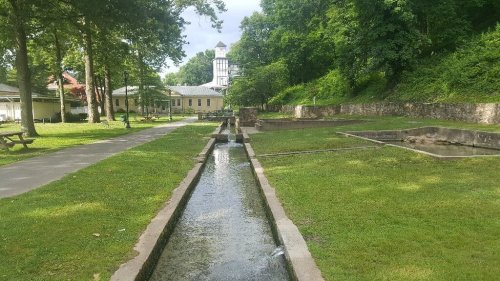 A serene park with a water channel, green grass, trees, and a historic building in the background under a cloudy sky.