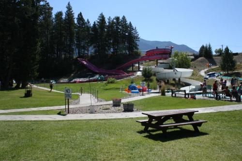 A sunny water park scene with slides, green grass, and picnic tables, surrounded by trees and mountains in the background.