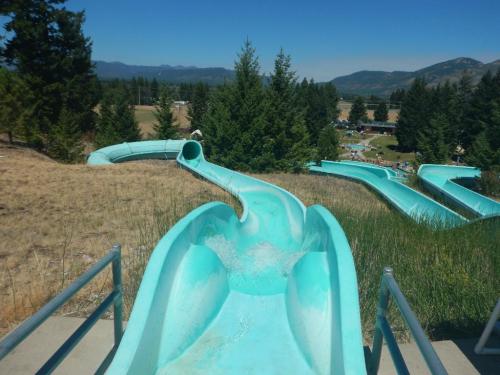 A turquoise water slide descends through a grassy area, surrounded by trees and mountains in the background.