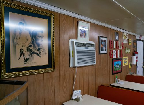 Interior of a diner featuring wooden walls, framed artwork, an air conditioner, and red signage reading "BILL'S GRILL."