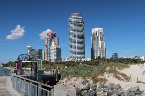 A view of modern skyscrapers along a beach, with a wooden pier and clear blue sky in the background.