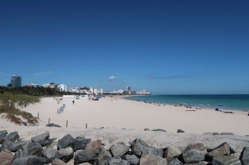 A wide view of a sandy beach with clear blue water, lined by buildings and palm trees under a bright blue sky.