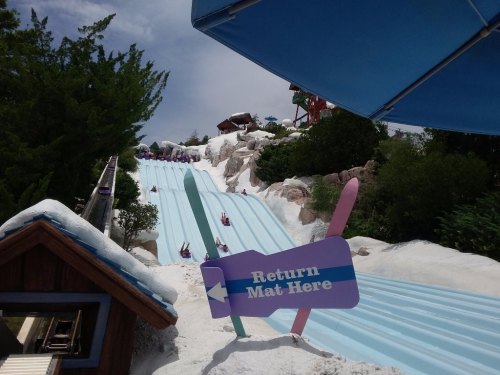 A water slide area with blue slides, surrounded by greenery, and a sign indicating where to return mats.