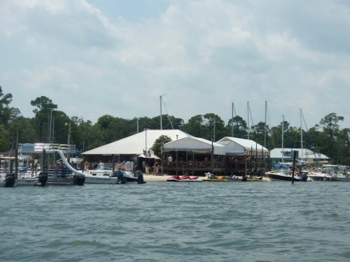 A waterfront scene featuring a restaurant, boats, and sailboats docked near a lush, green shoreline.