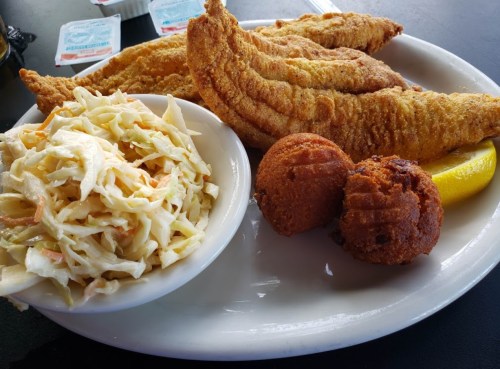 A plate of fried fish fillets with coleslaw, a cornbread muffin, and a lemon wedge.