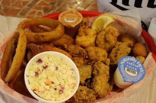 A basket of fried seafood, onion rings, coleslaw, and dipping sauces, garnished with a lemon wedge.