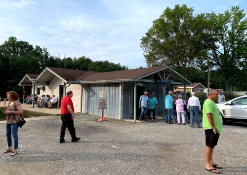 A small building with an "Open" sign, surrounded by people socializing outside on a gravel lot. Trees in the background.