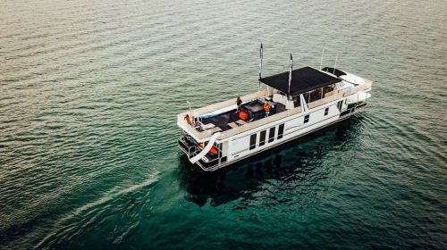 A houseboat floats on calm water, with two people on deck enjoying the serene surroundings.
