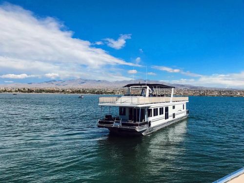 A houseboat glides across a calm lake under a blue sky with scattered clouds and distant mountains.