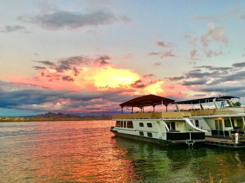 A houseboat docked on a lake at sunset, with vibrant clouds reflecting in the water.