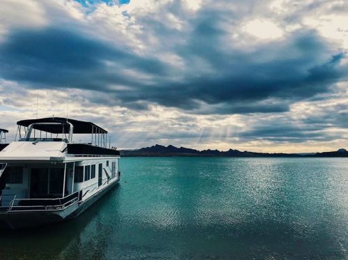 A houseboat docked on calm water under a cloudy sky, with mountains in the distance.