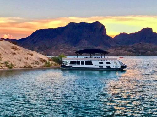 A houseboat anchored near a sandy shore, with mountains and a colorful sunset in the background.