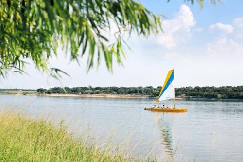 A colorful sailboat glides across a calm river, surrounded by lush greenery and a clear blue sky.