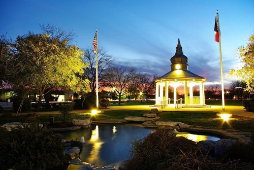 A gazebo illuminated at dusk, surrounded by trees and a pond, with flags in the background against a colorful sky.