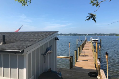 A wooden dock extends into a calm lake, with a small building and an American flag in the foreground under a clear blue sky.