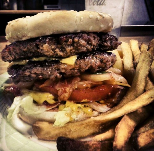 A towering burger with two patties, cheese, lettuce, tomato, and onions, served with a side of golden fries.