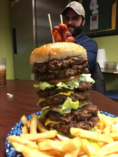 A towering burger with multiple patties, cheese, and lettuce, served with fries, with a man sitting in the background.