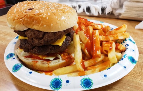 A double cheeseburger with lettuce and ketchup on a plate, accompanied by a side of French fries drizzled with ketchup.
