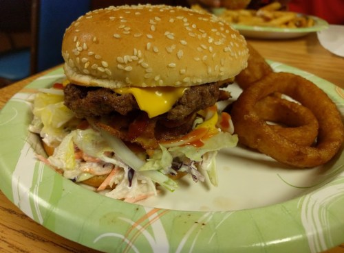 A sesame seed bun burger with cheese, bacon, and lettuce, served with onion rings on a paper plate.