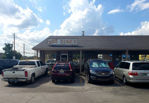 A diner with a sign reading "Little Diner," surrounded by parked cars under a partly cloudy sky.