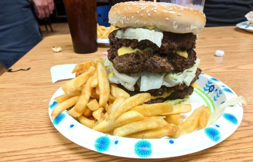 A towering burger with multiple patties, cheese, and lettuce, served with a side of golden French fries on a paper plate.