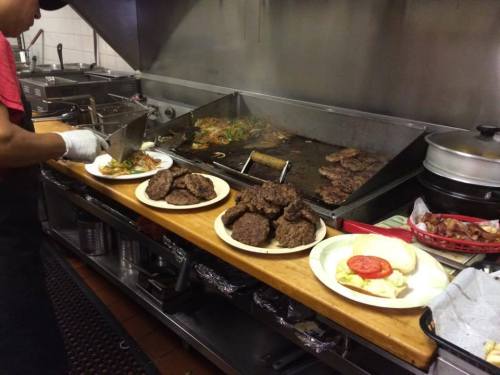 A chef prepares plates of food in a busy kitchen, with grilled patties and vegetables on the grill in the background.