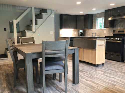 A modern kitchen with gray cabinets, a wooden dining table, and a staircase in the background. Bright and spacious.