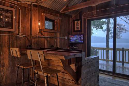 Cozy wooden interior with a bar, stools, and a view of a lake through large glass doors. Warm lighting enhances the ambiance.