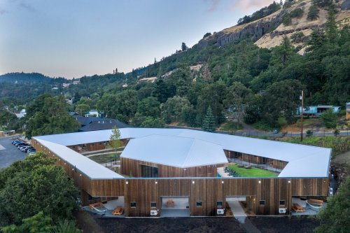 Aerial view of a modern, angular wooden building surrounded by trees and hills, with a parking area nearby.