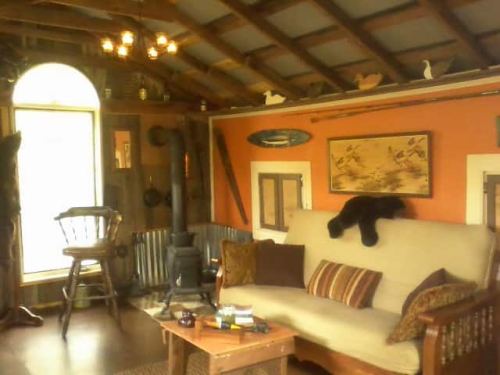 Cozy cabin interior with a sofa, wooden decor, a stove, and rustic furnishings under a sloped ceiling.