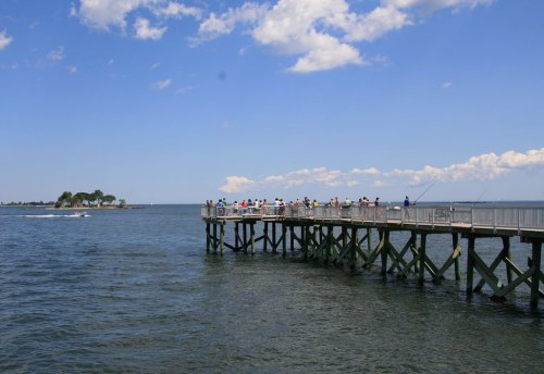 A wooden pier extends over calm water, with people fishing and a boat passing by under a blue sky with clouds.