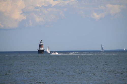 A lighthouse stands in the water with sailboats and a motorboat nearby under a blue sky with clouds.