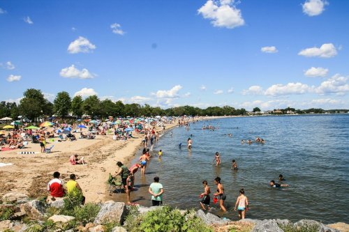 A busy beach scene with people swimming, sunbathing, and enjoying the sunny day under a blue sky with fluffy clouds.