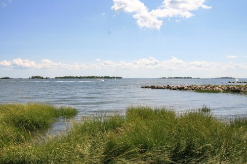 A serene view of a coastal landscape with grassy foreground, calm water, and distant islands under a blue sky with clouds.
