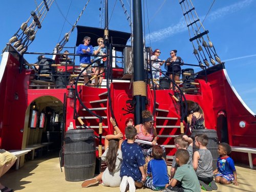 A group of children sits on the deck of a red pirate ship while adults stand above, enjoying a sunny day.