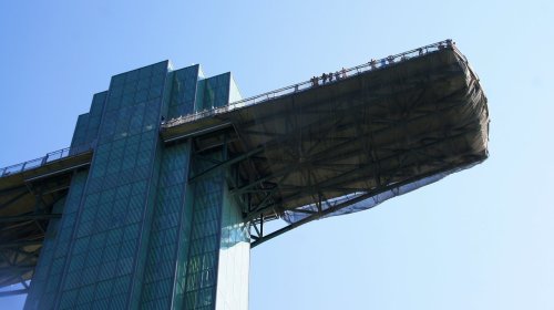 A tall, modern building with a large, cantilevered platform extending from the top against a clear blue sky.