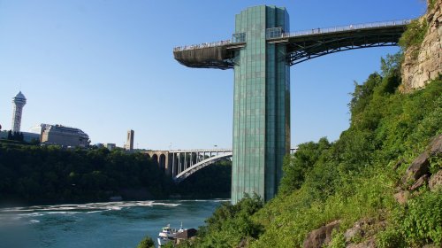 A glass observation tower overlooks a river, with a bridge and buildings in the background under a clear blue sky.