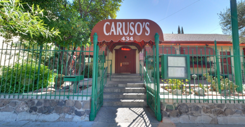 Entrance to Caruso's restaurant, featuring a red awning and green fence, surrounded by plants and cacti.