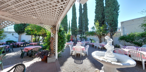 A sunny outdoor dining area with tables and chairs, surrounded by greenery and a fountain.